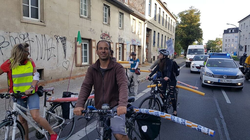 Bei der Poolnoodle-Tour: Eine Gruppe von Menschen auf Fahrrädern steht auf einer Straße. Auf den Gepäckträgern haben sie Poolnoodlen. EIn VCDler lacht in die Kamera.