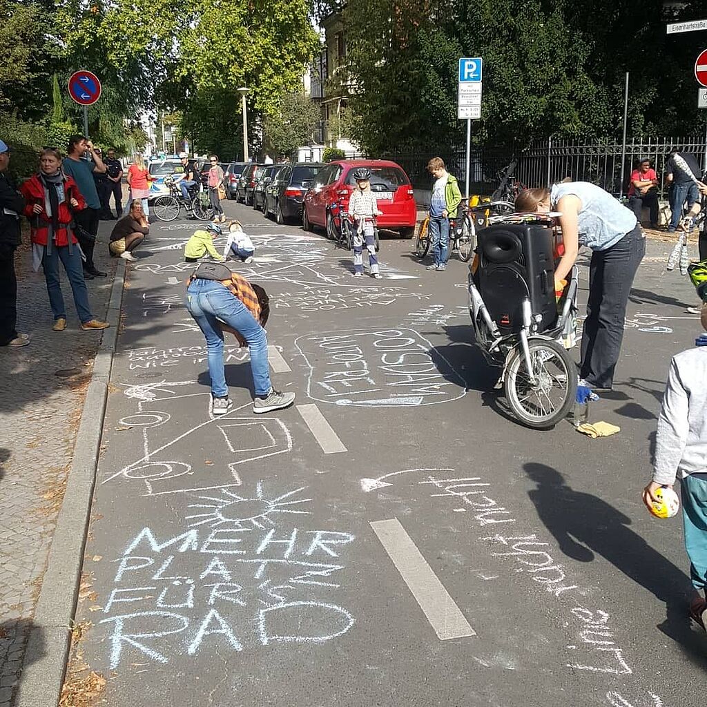 Bei der Kidical Mass: Eine Straße ist mit bunten Kreidemalereien bedekct, zum Beispiel sthet dort "Mehr Platz fürs Rad". Kinder und Erwachsene malen noch mehr.