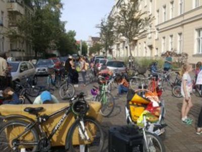 Bei der Kidical Mass: Viele Menschen und Fahrräder stehen weit vereteilt auf einer Straße. Die Menschen malen mit Kreide, spielen, sind in Gesprächen. Mitten auf der Straße steht außerdem ein Sofa.