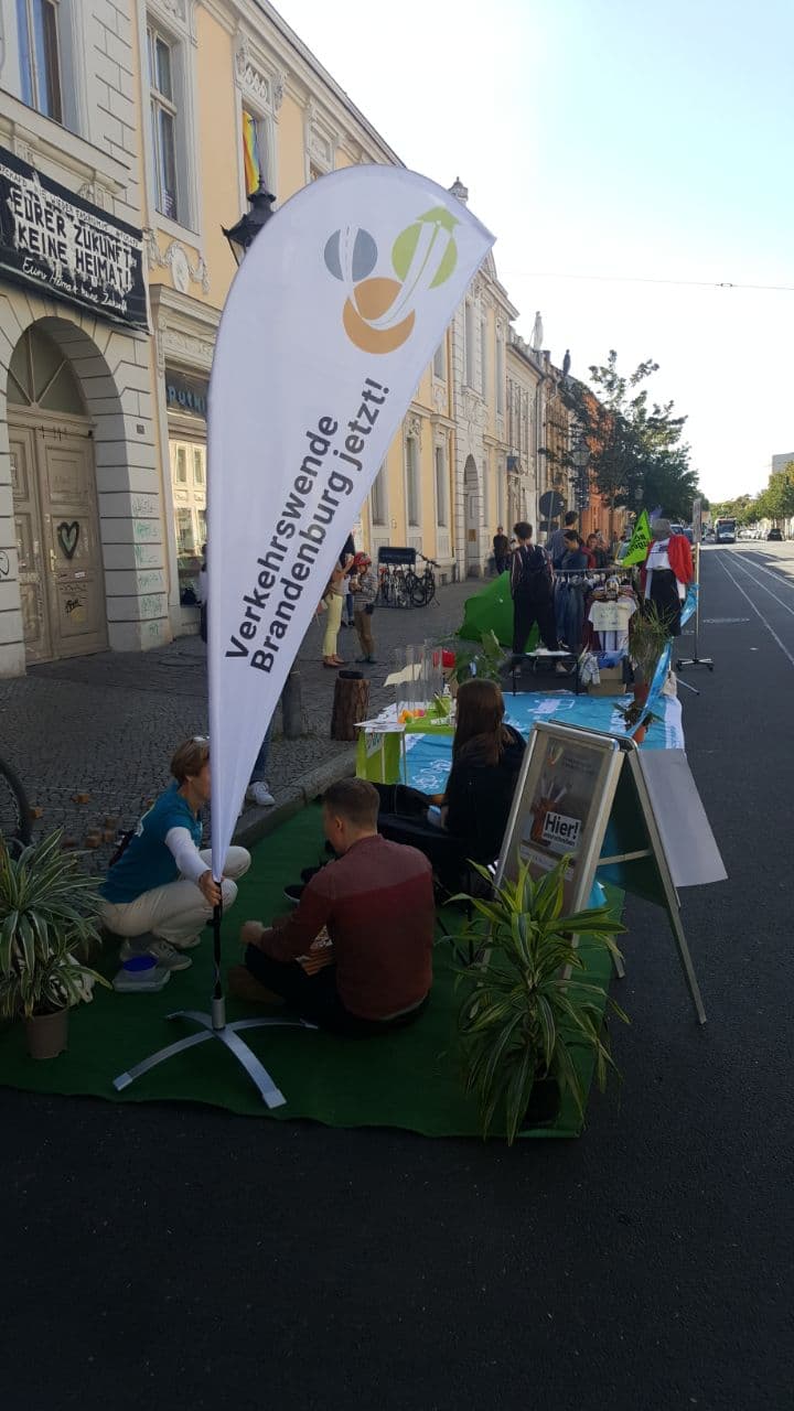 Viele Menschen sitzen auf Plätzen, die sonst zum Parken genutzt werden. Auf dem Boden liegt grüner Stoff, es wurden viele Pflanzen, Schilder und Spiele aufgestellt. Im Vordergrund ist eine Beachflag der Volskinitiative "Verkehrswende Brandenburg jetzt!" zu sehen.